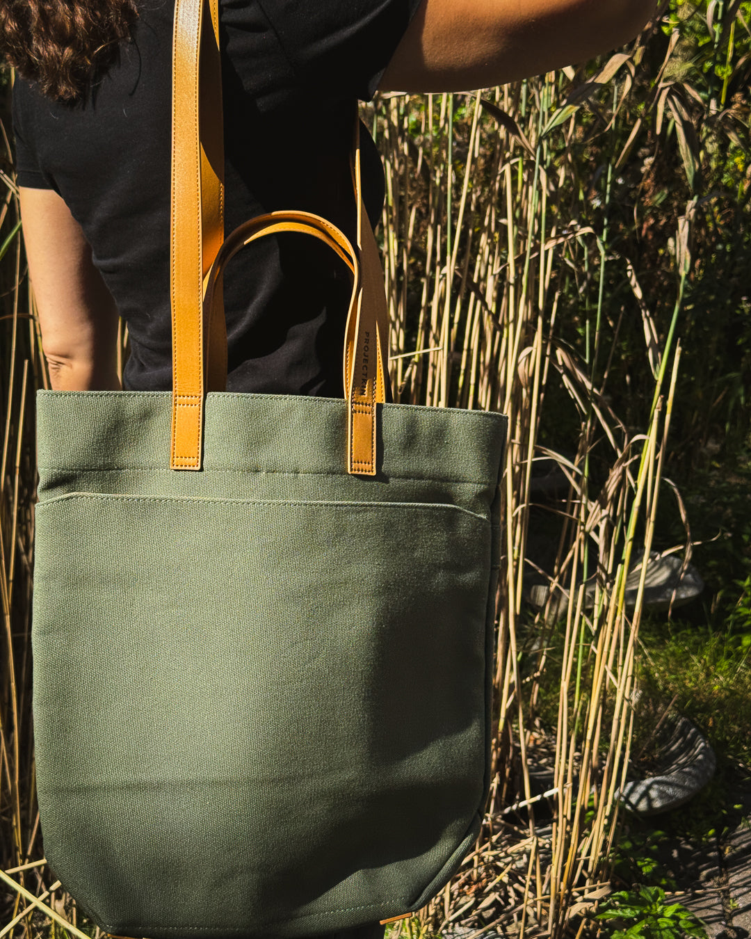 An olive green canvas tote bag with tan vegan-leather handles, carried by a person standing in tall dry grass outdoors.