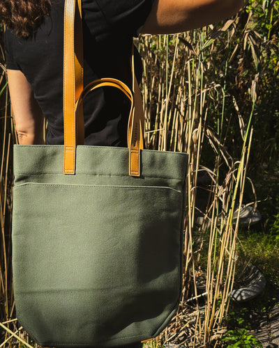 An olive green canvas tote bag with tan vegan-leather handles, carried by a person standing in tall dry grass outdoors.