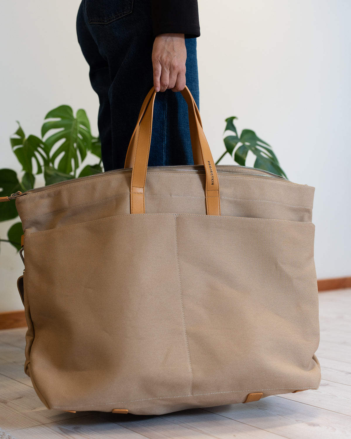 A large beige canvas bag with tan vegan-leather handles, held by a person wearing dark jeans in a bright room with a potted green plant in the background.