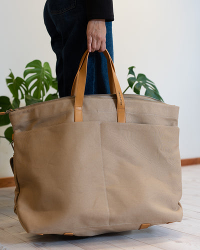 A large beige canvas bag with tan vegan-leather handles, held by a person wearing dark jeans in a bright room with a potted green plant in the background.
