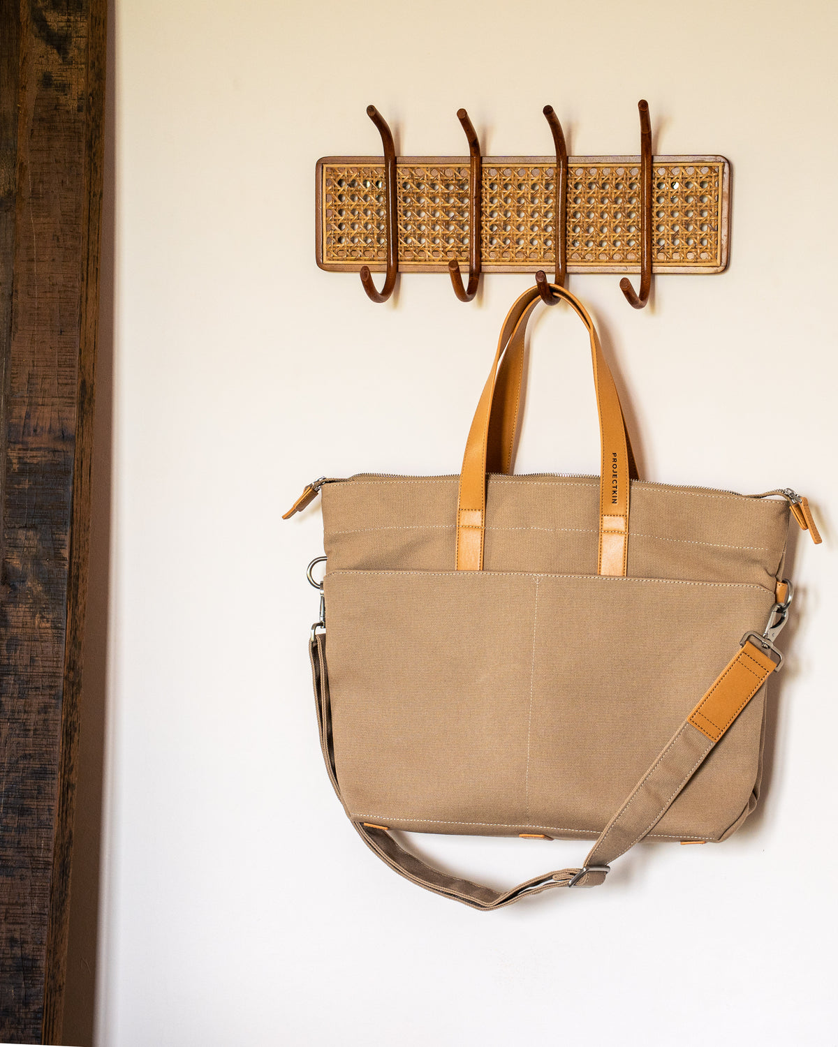 A beige canvas tote bag with tan vegan leather handles and a matching shoulder strap hangs from a woven wall hook rack against a white wall.